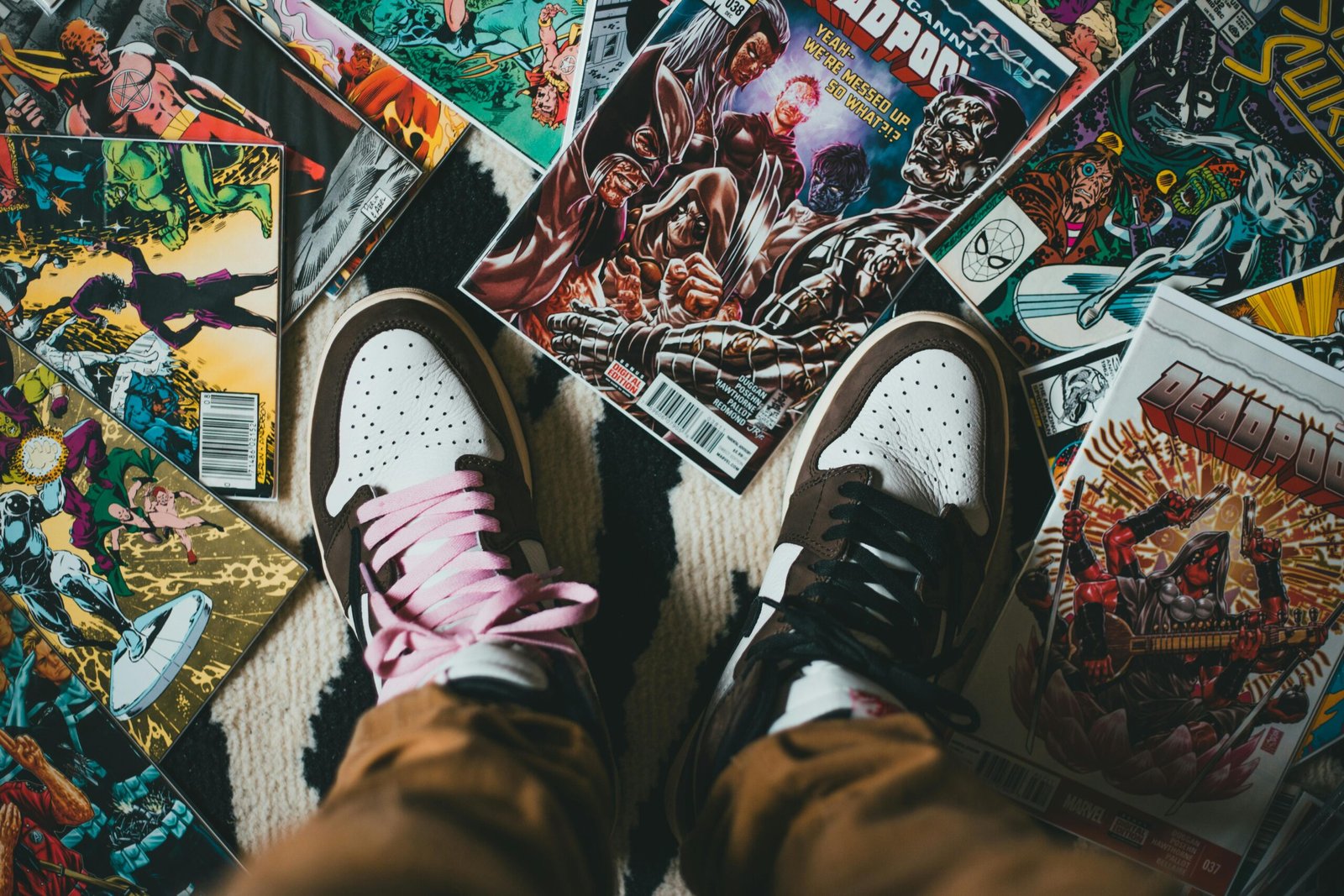 From above crop person wearing brown trousers and sneakers standing on pile of collection of comics magazines with colorful illustrations on cover